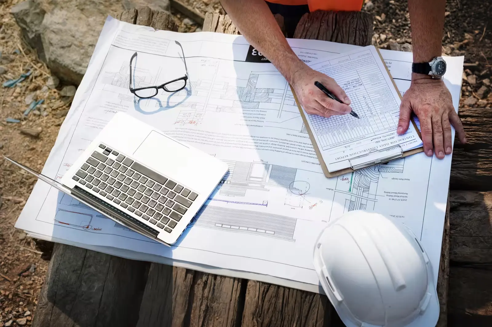 mãos masculinas escrevendo numa prancheta por cima de uma grande planta de projeto junto com capacete, notebook e um pár de óculos, todos sobre uma mesa de troncos ao ar livre